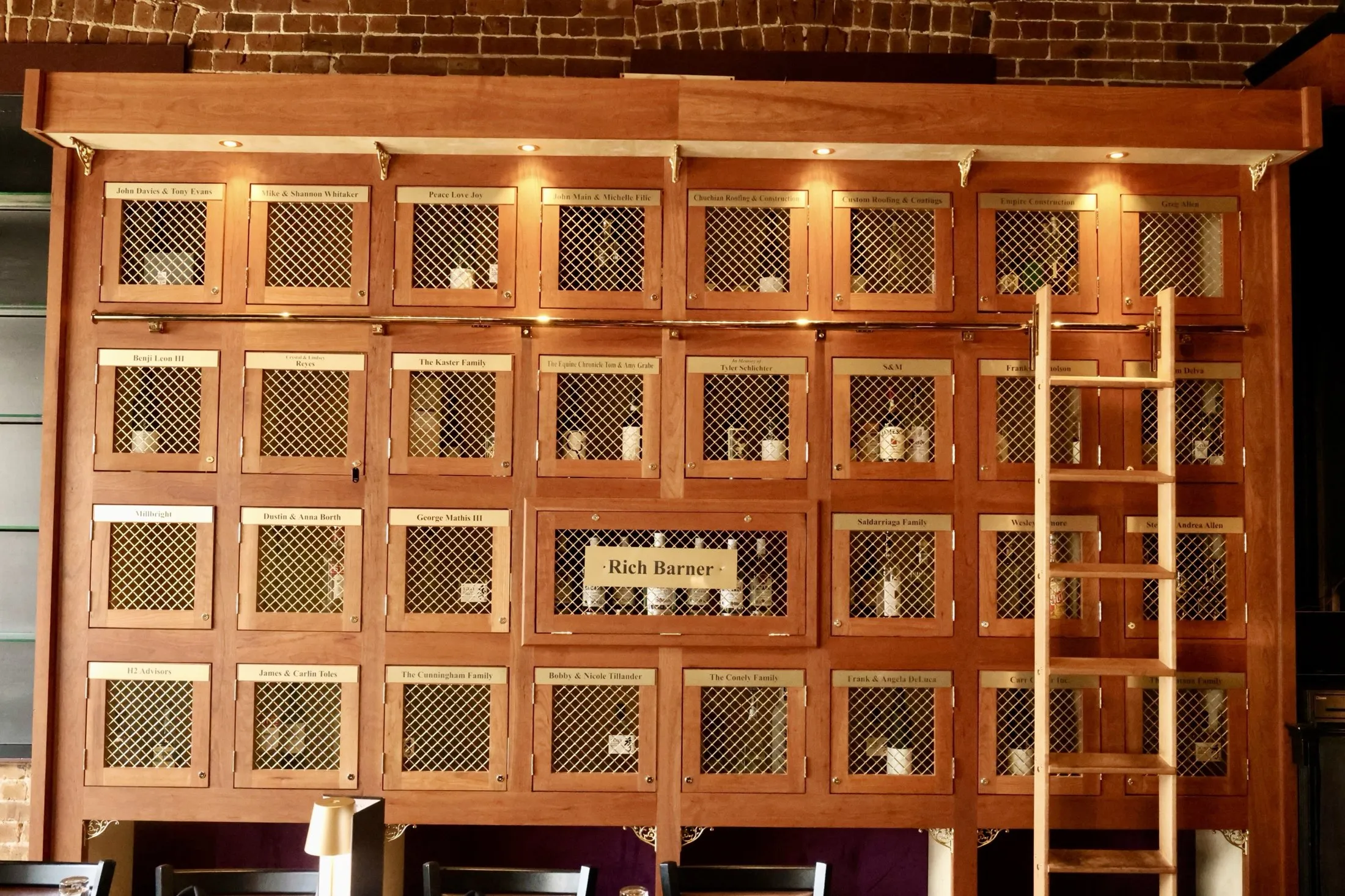 The bottle locker wall at Katya Vineyards in downtown Ocala: cherry-wood lockers with brass mesh doors and engraved member nameplates, lit beneath the brick ceiling, with a rolling library ladder